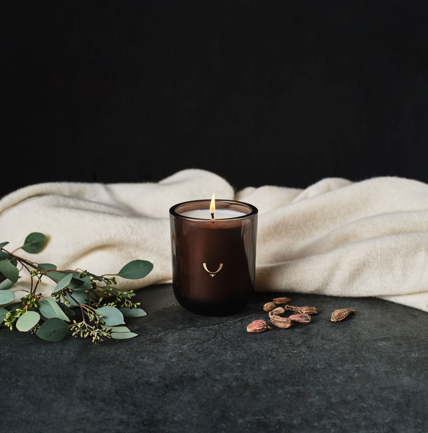 Lit candle in a setting with a white blanket, leaves and cardamon pods